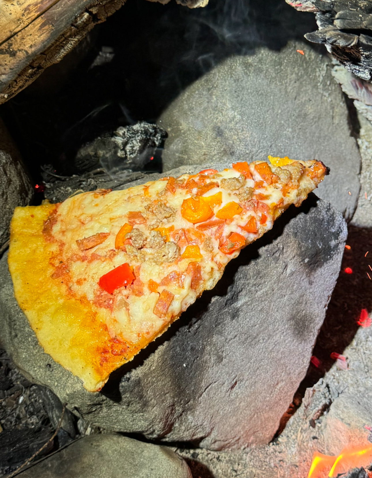 Slice of pizza with red peppers and sausage on a stone near a campfire. Brightly lit, contrasting with dark, smoky background.